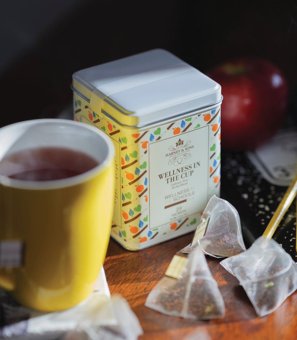 Tea canister labeled Wellness in the Cup with a yellow mug and tea bags on a wooden surface.