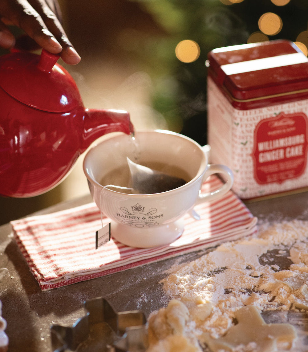 Person pouring tea from a red teapot into a white cup on a wooden surface with a ginger cake box in the background.
