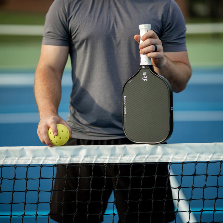 Person holding a pickleball paddle and ball on a tennis court.