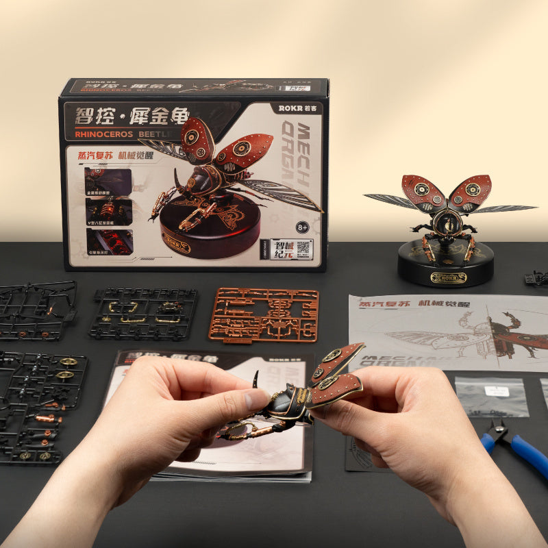 Person assembling a mechanical model of a butterfly with packaging and tools on a table.