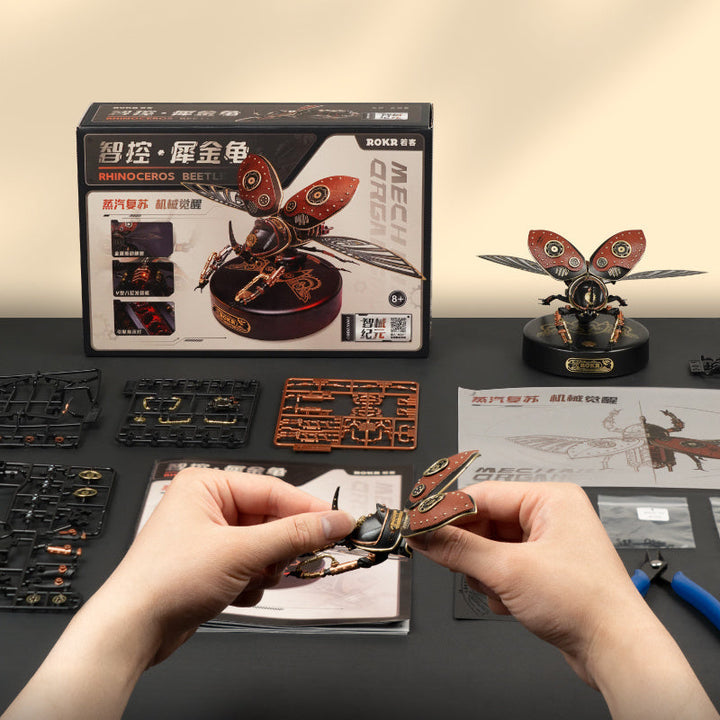 Person assembling a mechanical model of a butterfly with packaging and tools on a table.
