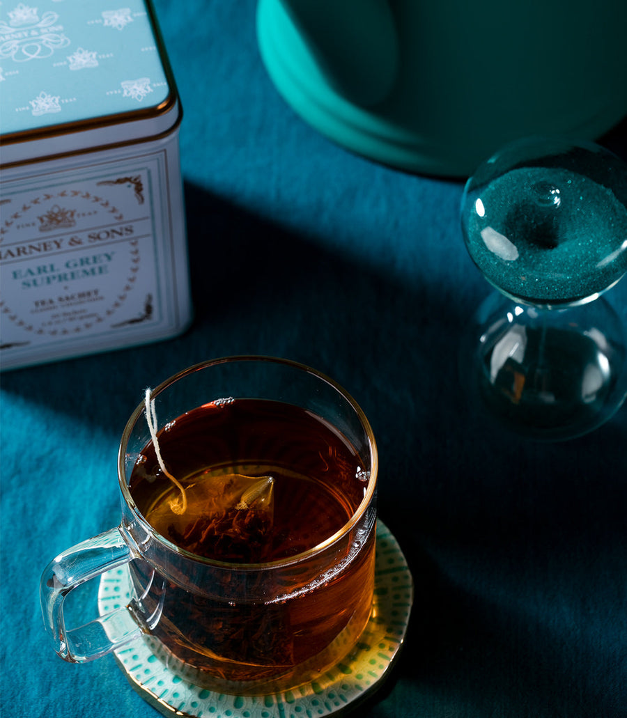 Glass cup of tea with a teabag, next to a tin labeled Barney and Sons Earl Grey Supreme on a teal surface.