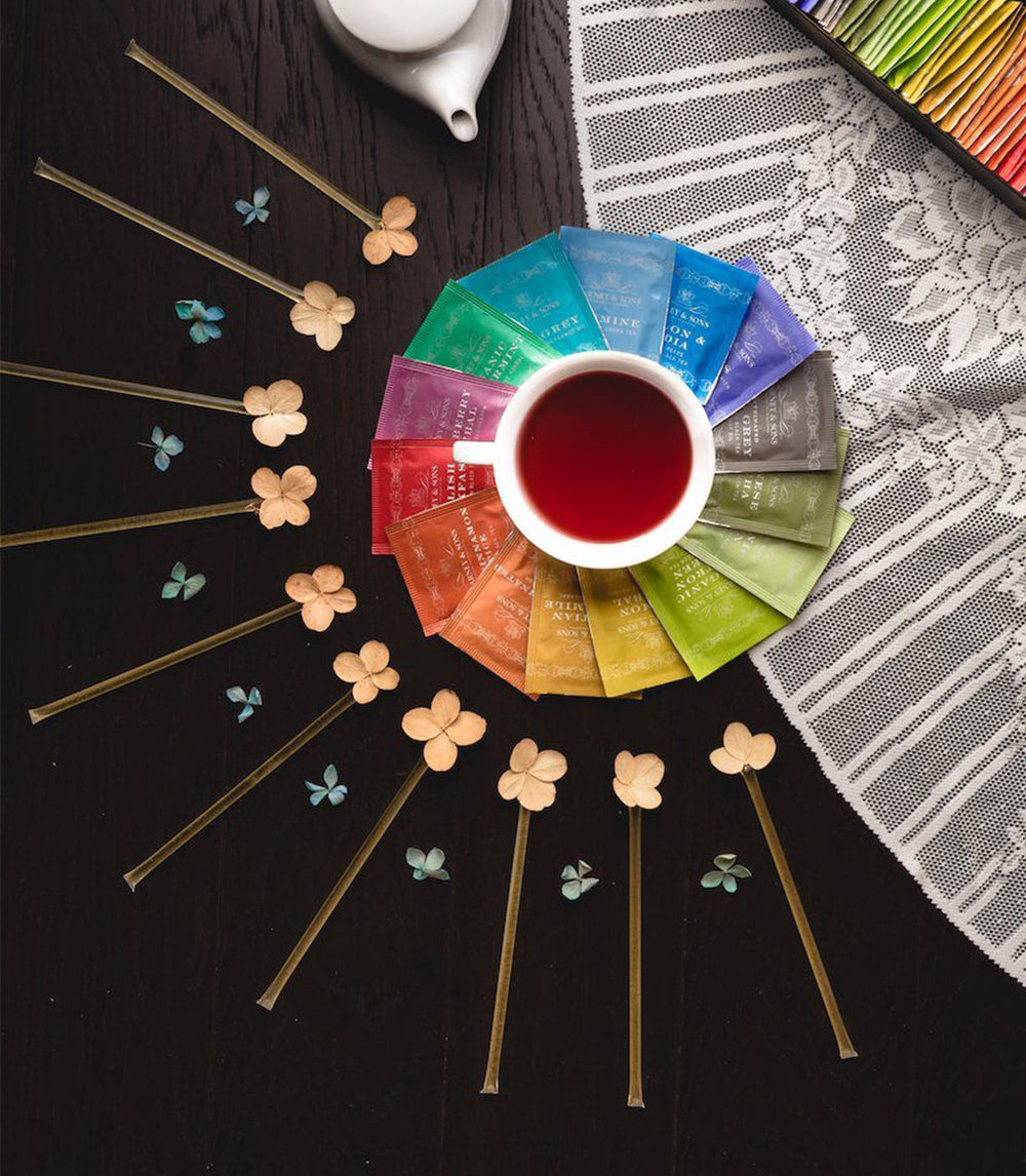 Tea bags arranged in a color wheel with a cup of tea in the center on a dark surface.