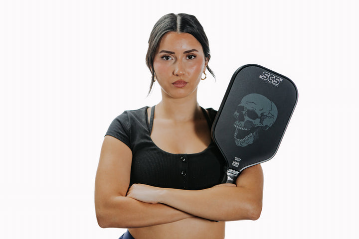 Woman holding a pickleball paddle with a skull design on a white background.