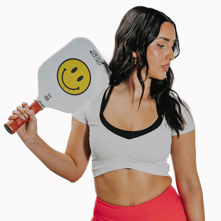 Woman holding a pickleball paddle with a smiley face design on a white background.