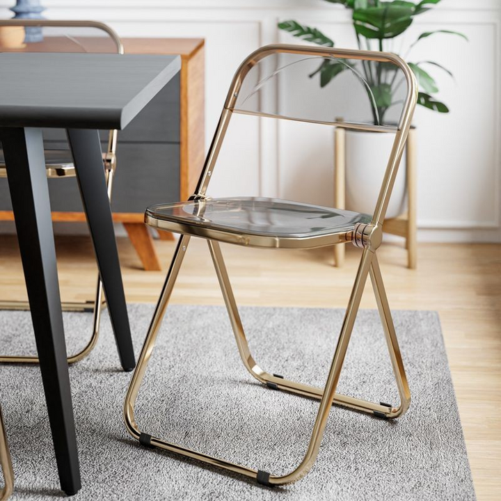 LeisureMod Lawrence Acrylic Folding Chair with Gold Metal Frame Transparent Black at Table.